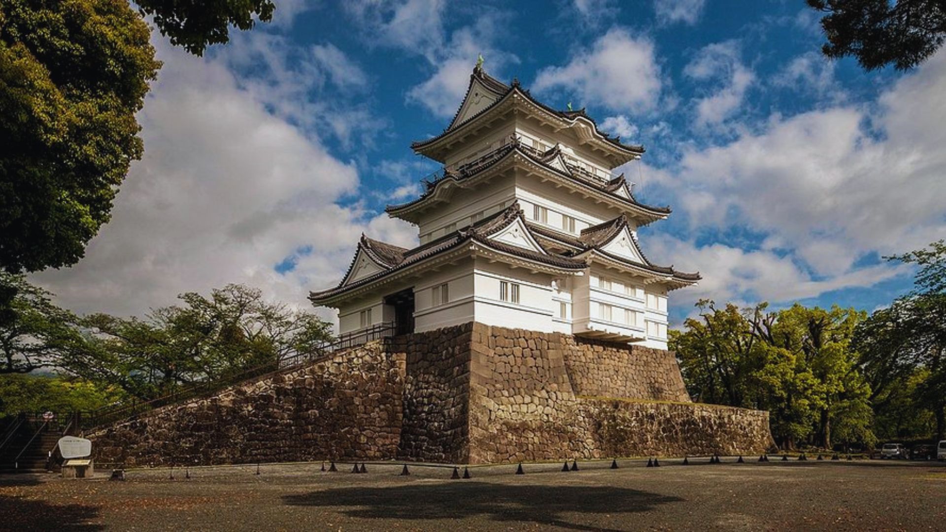 A traditional Japanese castle with white walls and layered roofs stands on a stone base under a blue sky with clouds, surrounded by lush green trees.