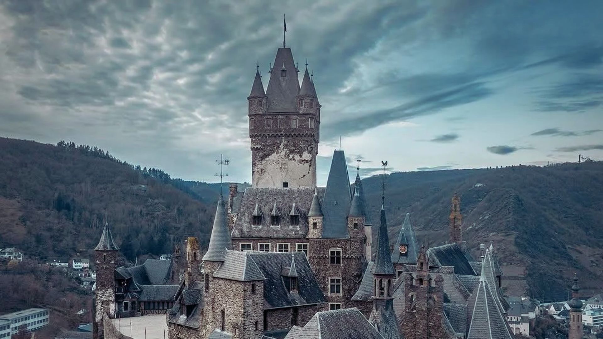 A medieval castle with tall spires and a central tower stands against a backdrop of rolling hills and a cloudy sky, creating a mysterious and historic atmosphere.