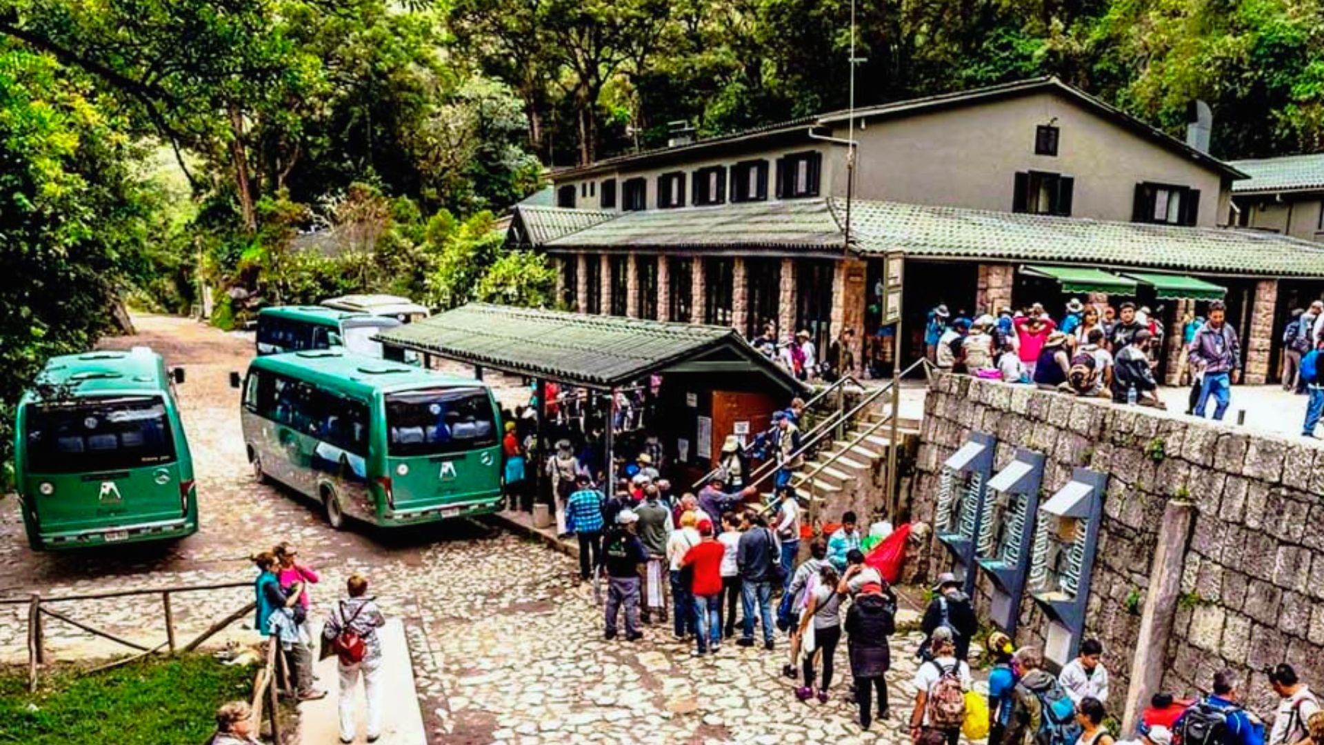 A busy tourism hub features green buses parked near a rustic building surrounded by lush trees. Crowds of visitors gather, creating a lively atmosphere.