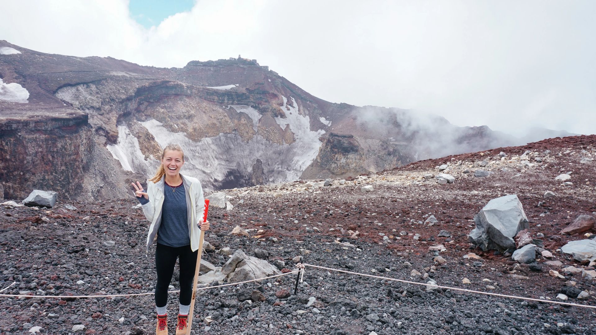 A woman smiles and gives a peace sign while standing on a rocky, volcanic landscape. Mist and a steep crater wall create a dramatic backdrop.