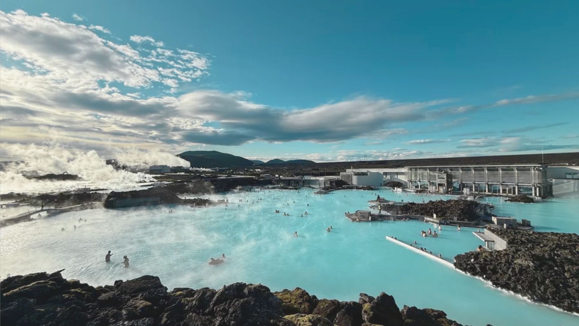 A serene geothermal lagoon with milky blue water, surrounded by black volcanic rocks. Bathers enjoy the warm water under a partly cloudy sky.