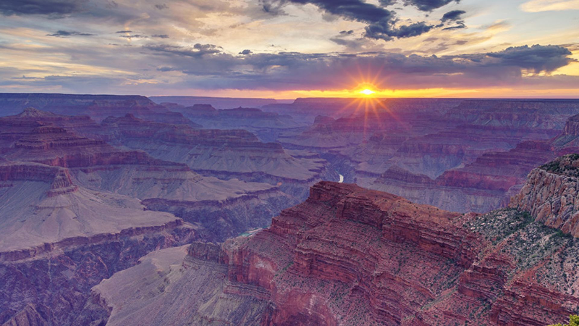Sunset over the Grand Canyon, with vibrant oranges and purples illuminating layered rock formations and casting dramatic shadows across the vast landscape.