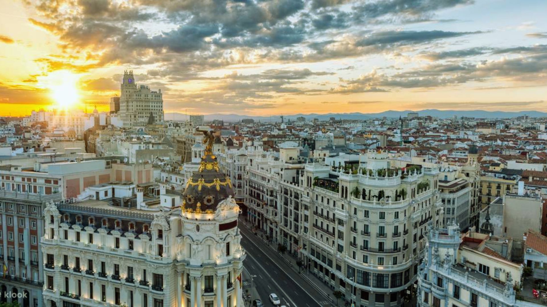 Aerial view of Madrid at sunset, highlighting historic buildings and the iconic Metropolis dome. The sky is vibrant with warm oranges and soft clouds.