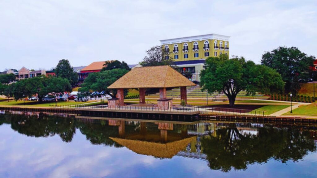A gazebo on the water in front of a building, surrounded by calm water and greenery.