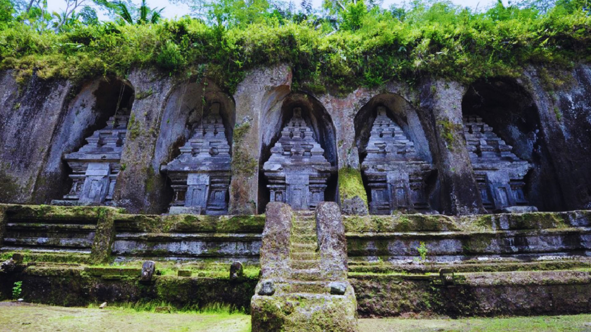 Ancient stone shrines carved into a mossy cliff, framed by lush greenery. Five ornate alcoves house temple structures, evoking a serene, historical ambiance.