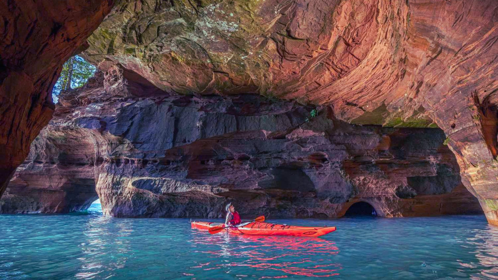 A person kayaks through vibrant blue waters inside a grand, rocky cave with textured red and brown walls, evoking a sense of adventure and tranquility.