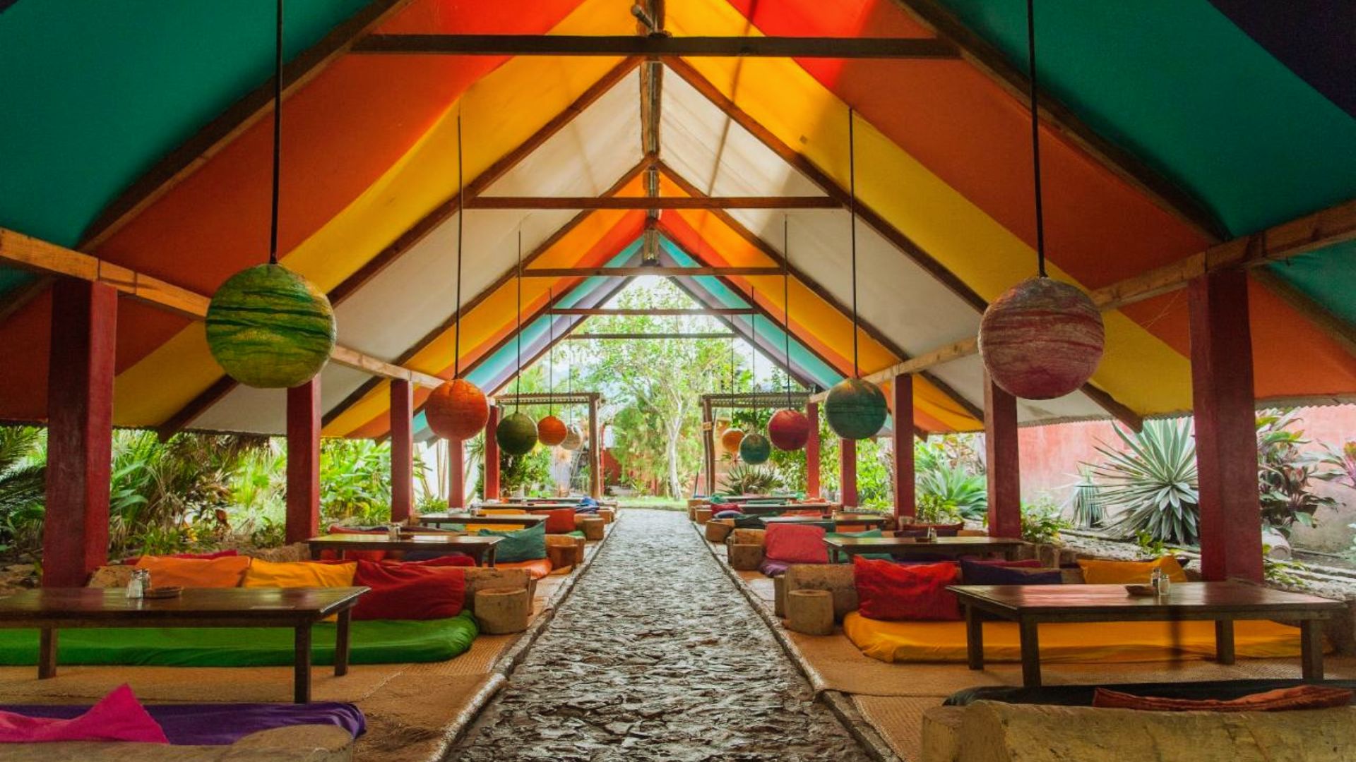 Colorful open-air restaurant with a vaulted striped roof and vibrant hanging lanterns. Cushioned seating on either side of a stone path, exuding a relaxed, inviting feel.