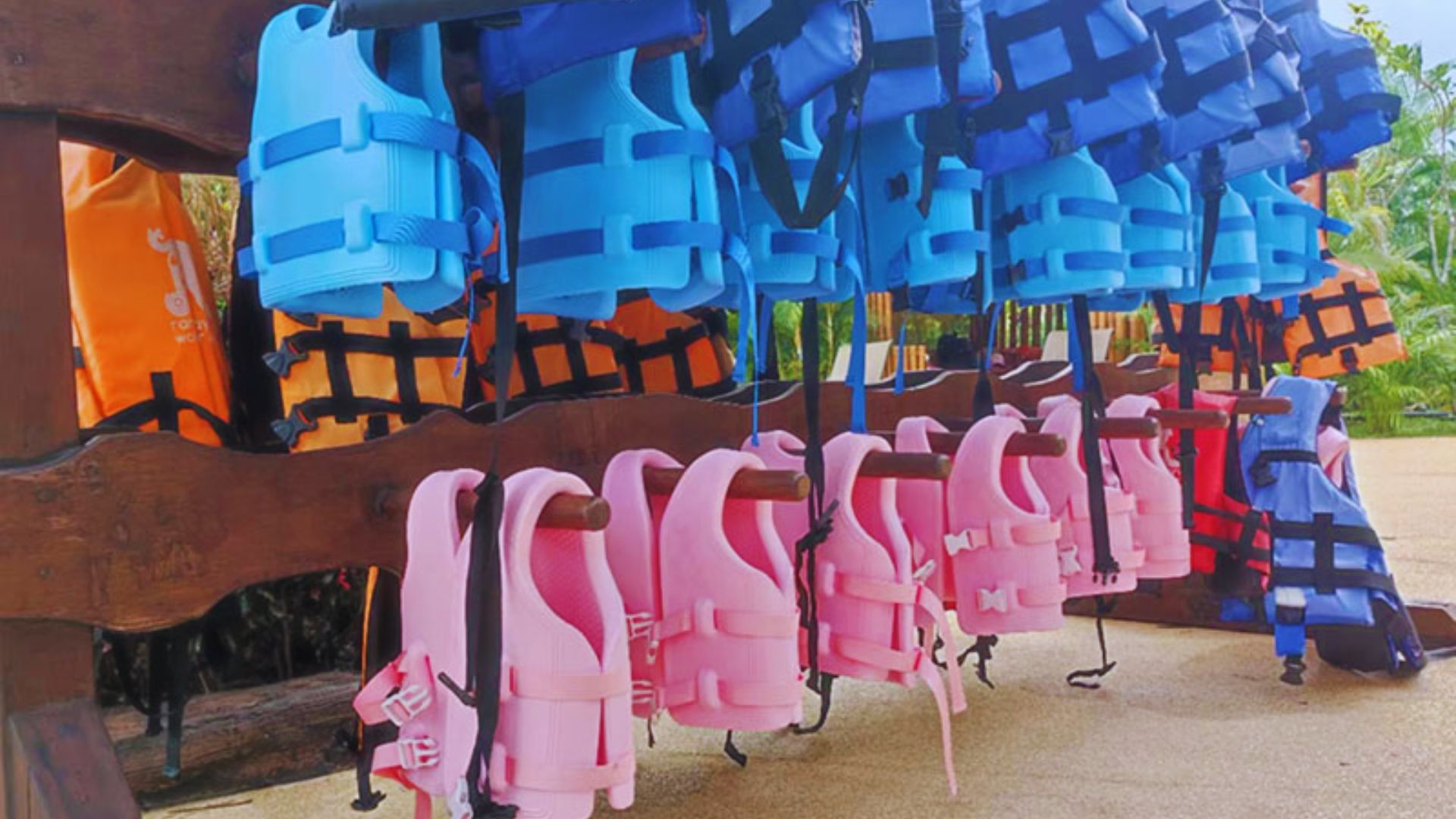 A row of colorful life jackets, including blue, pink, orange, and red, hang from a wooden rack. The setting is outdoors, with greenery in the background.