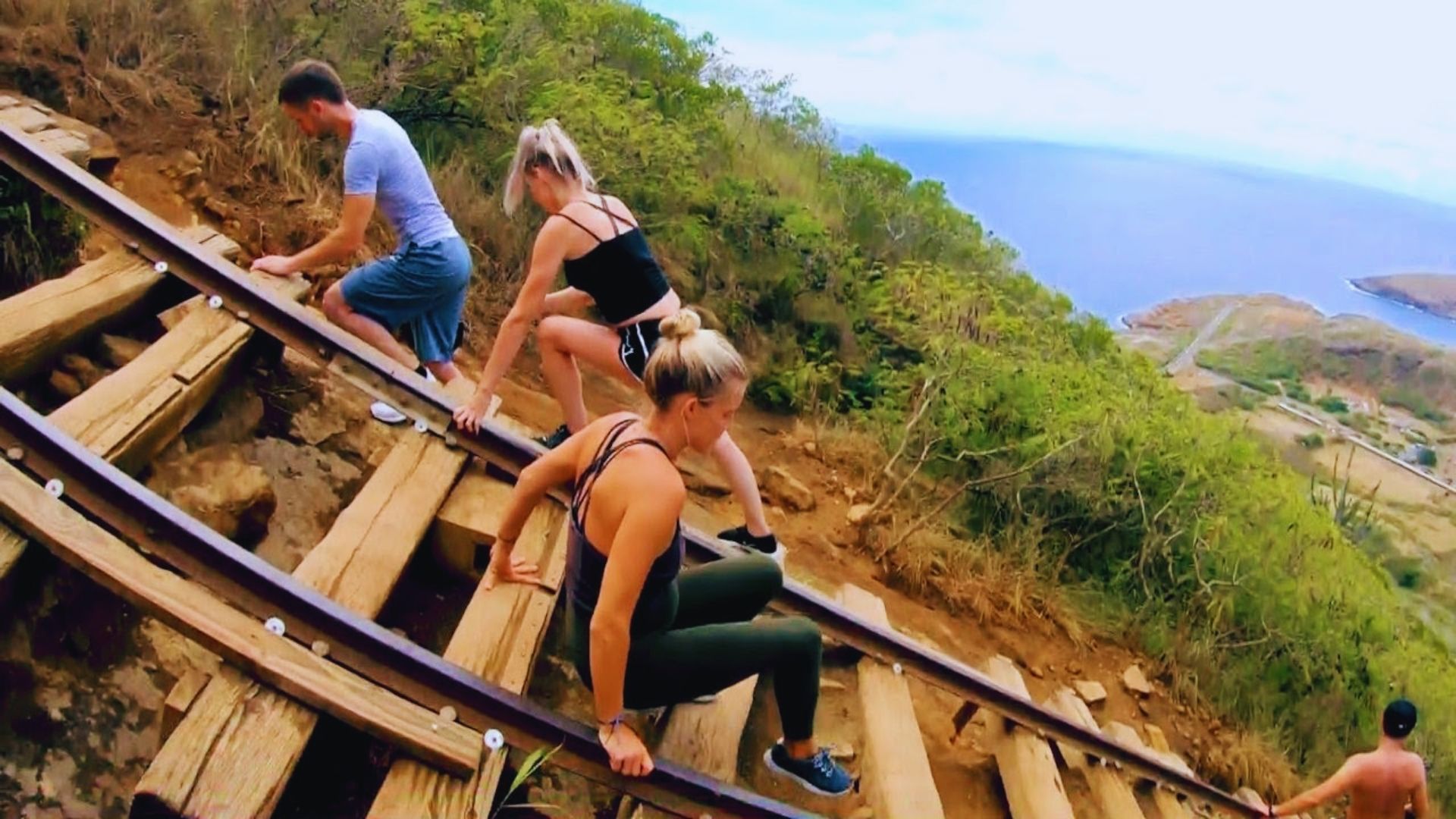 Three people carefully descend a steep wooden trail with rail tracks on a lush hillside overlooking the ocean, conveying a sense of adventure and concentration.