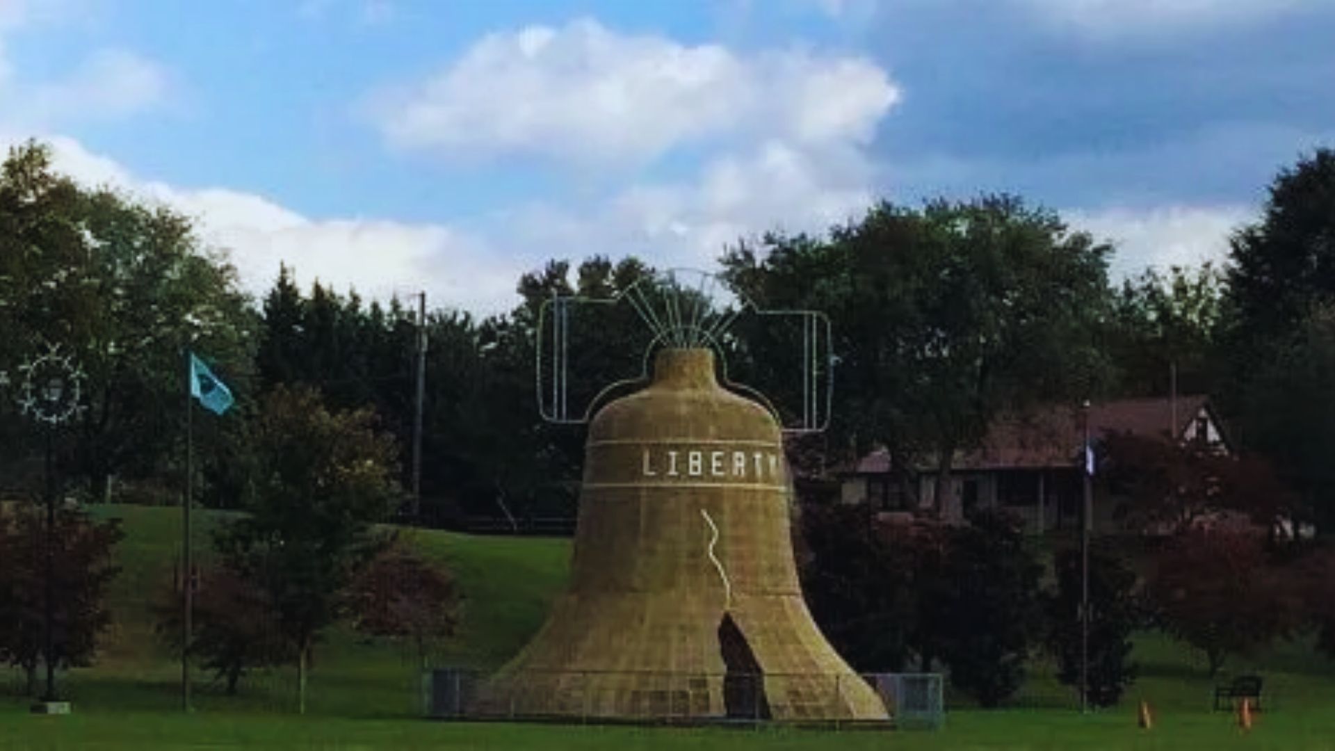 A large replica of the Liberty Bell, featuring "LIBERTY" and a crack, stands on grassy terrain with trees and a few flags in the background under a partly cloudy sky.