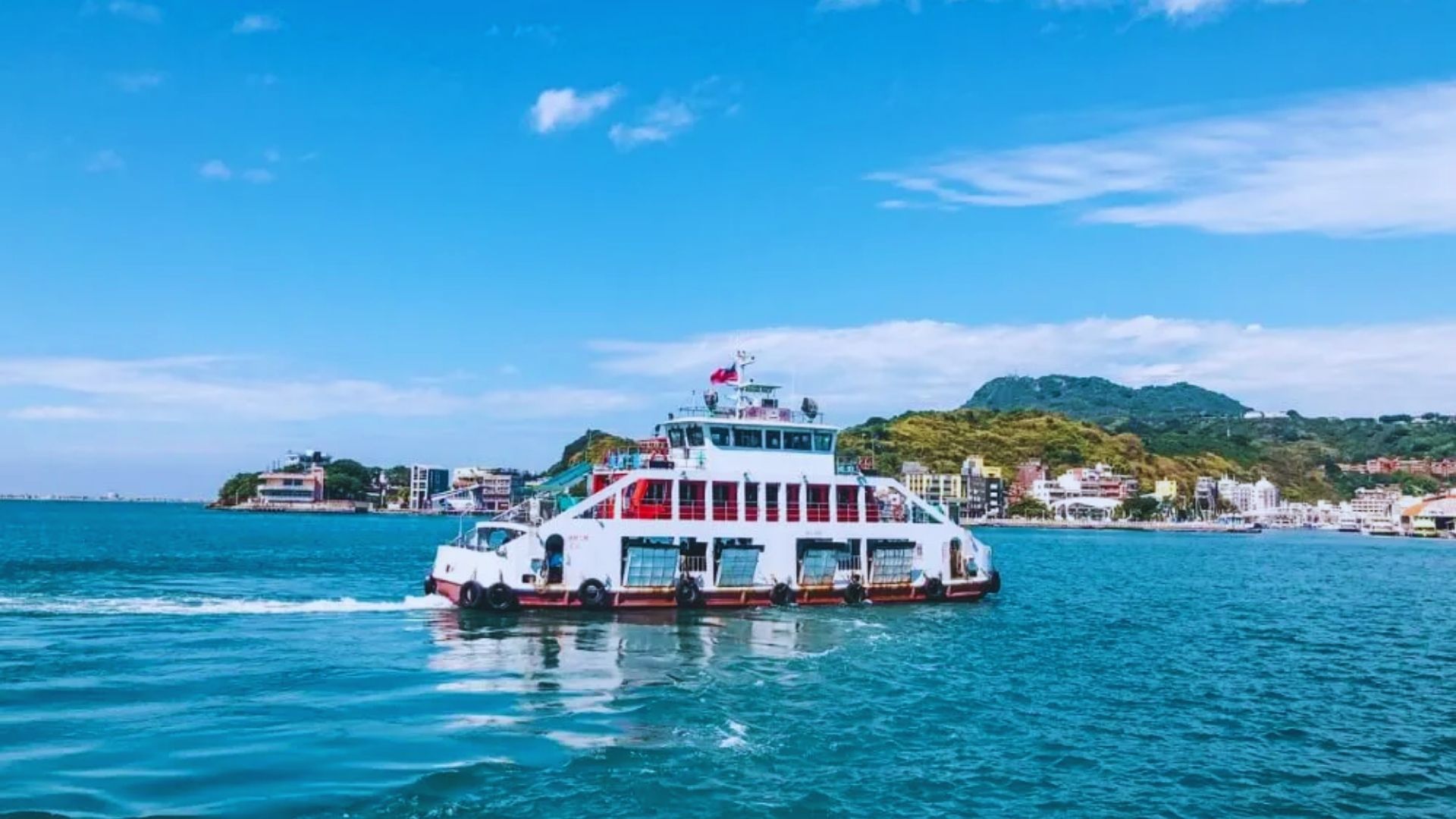 A white ferry with multiple decks crosses blue water on a sunny day. In the background, there are coastal buildings and green hills under a clear sky.