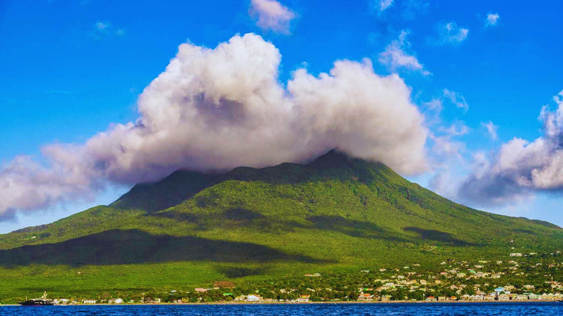 Lush green mountain under a blue sky with white clouds covering the peak. Small town at the base, and a calm blue sea in the foreground. Serene atmosphere.