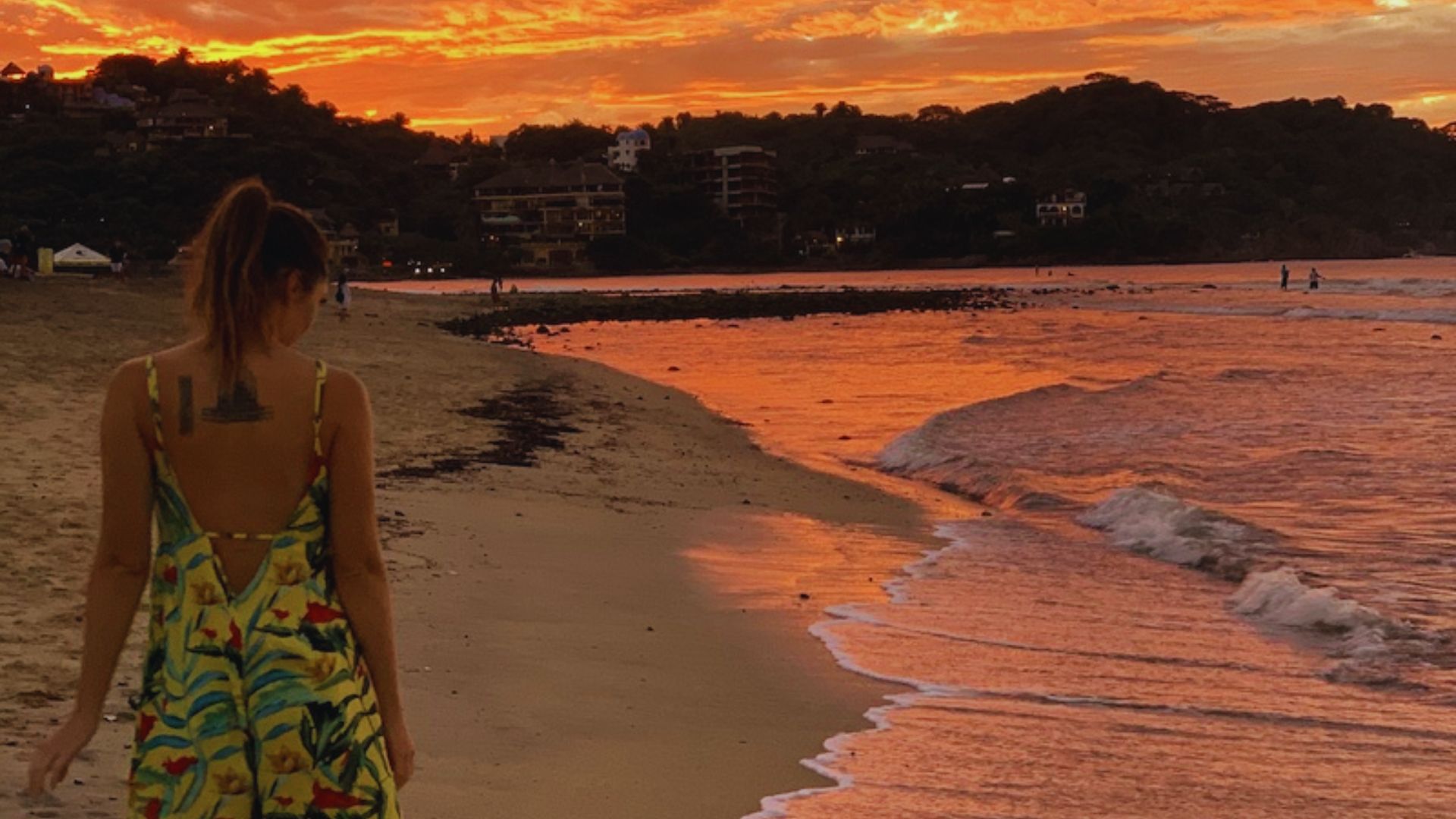 A woman in a colorful sundress walks along a beach at sunset. The sky and sea glow with vibrant orange hues, creating a peaceful and serene atmosphere.