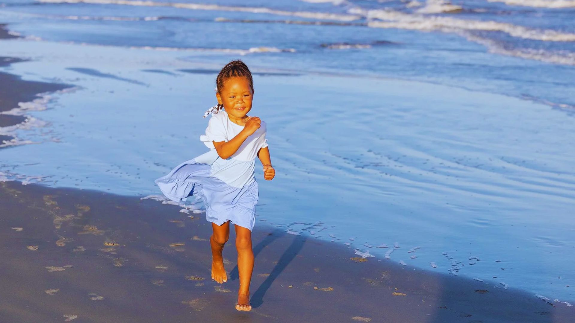 A joyful child in a light blue dress runs barefoot along a sunny beach. The ocean waves gently lap at the shore, capturing a carefree, happy moment.