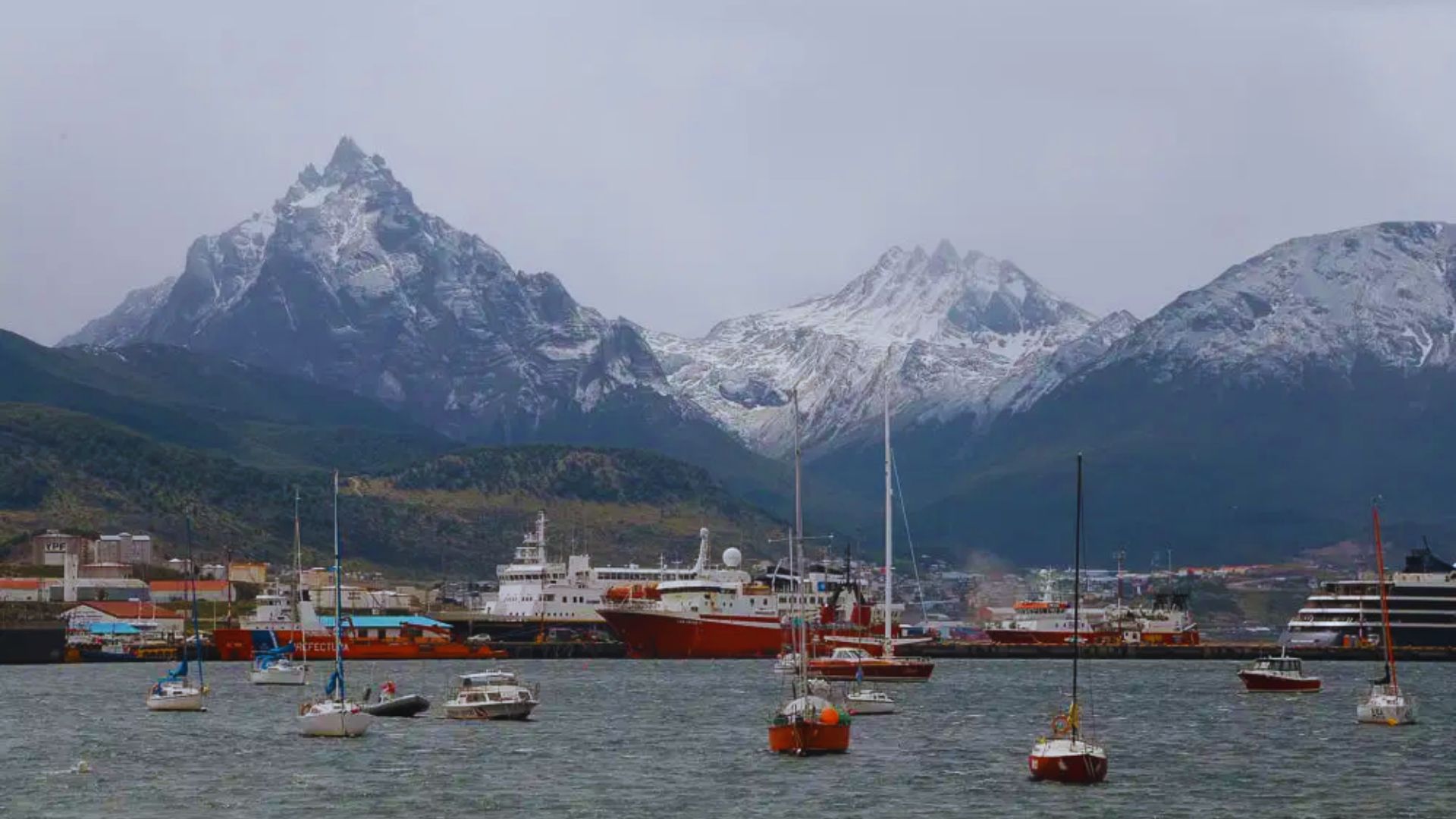 Snow-capped mountains tower over a harbor with sailboats and a large red ship. Overcast sky and calm waters convey a serene, chilly atmosphere.