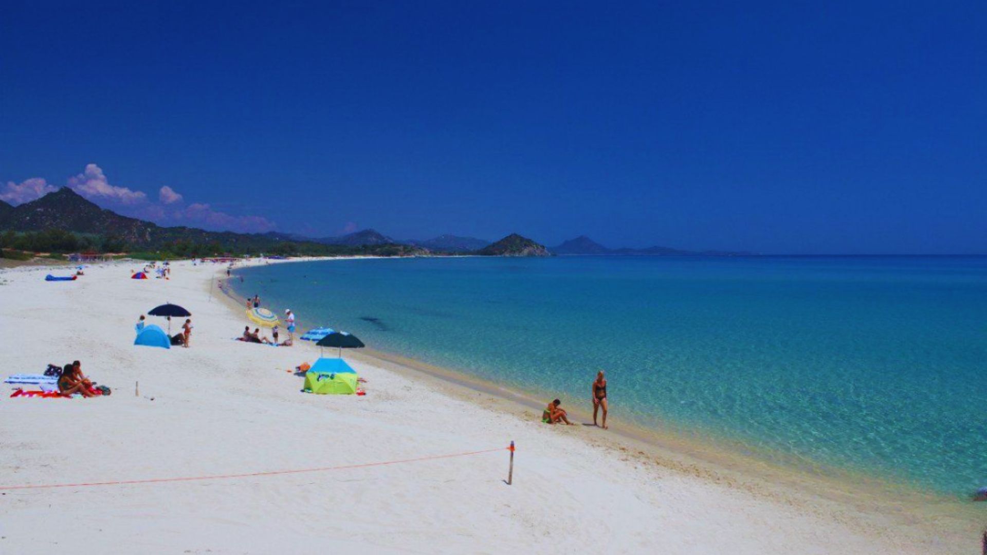 A serene beach scene with white sand, turquoise water, and a clear blue sky. Sunbathers and colorful umbrellas dot the shore, with distant mountains.
