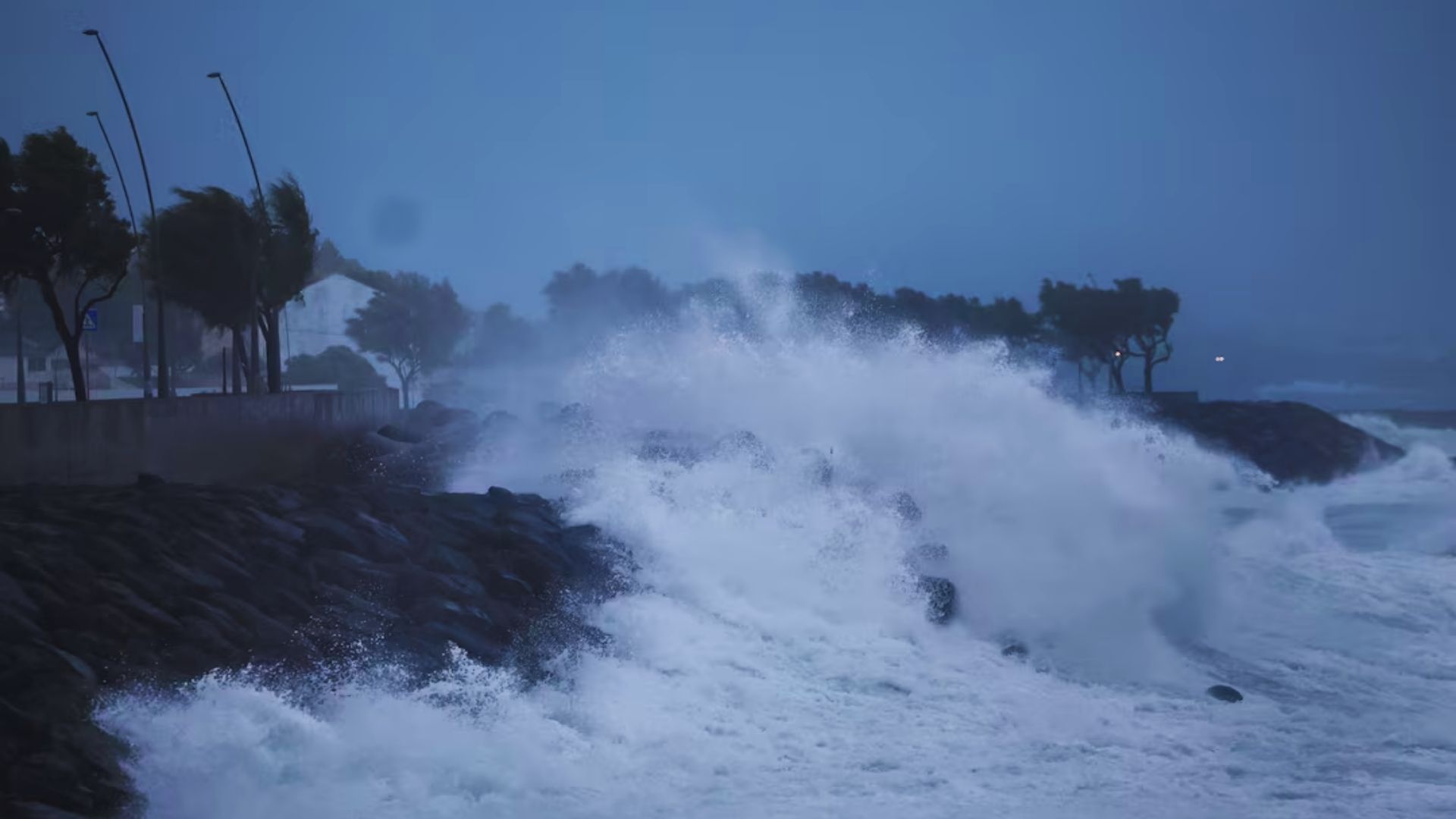 A large wave crashes dramatically over rocky formations near a sandy beach, showcasing the power of the ocean.