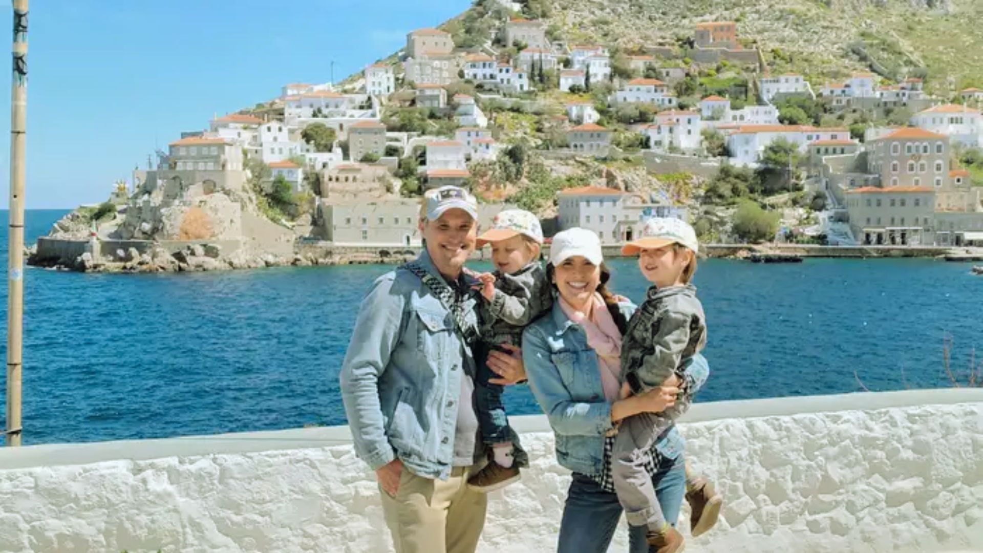 A smiling family of four poses by the sea, with parents holding children. They stand in front of a scenic hillside village with white houses under a clear blue sky.