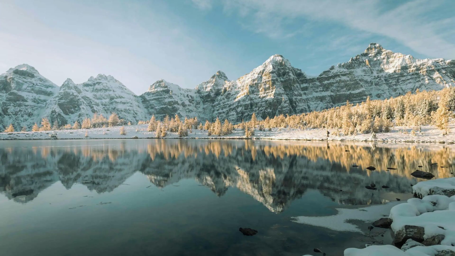 Snow-capped mountains reflecting in a calm lake under a clear blue sky; golden trees line the shore, creating a serene, peaceful atmosphere.