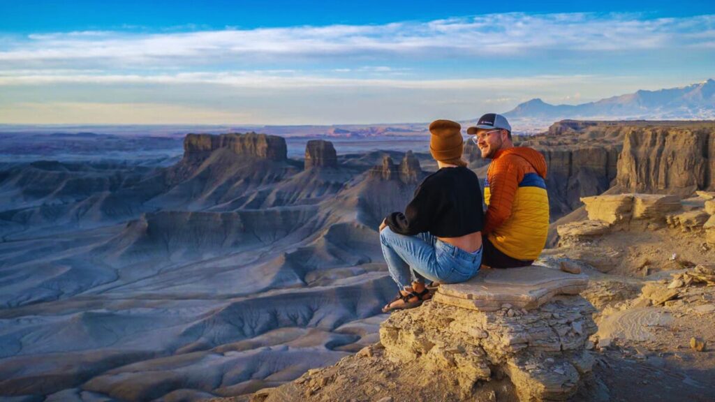 Two people sit on a rocky ledge, overlooking a vast desert landscape with dramatic mesas at sunset. They appear relaxed, enjoying the breathtaking view.