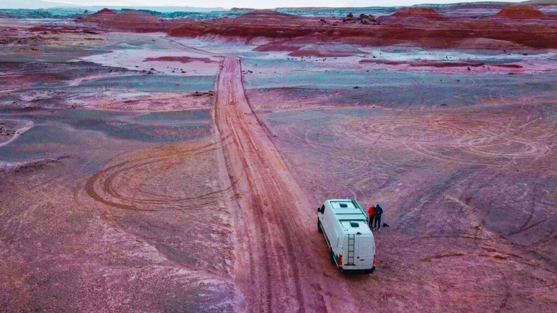 A white van on a reddish-brown dirt road in a vast, barren desert landscape. Two people stand by the van under a cloudy sky, evoking exploration and solitude.