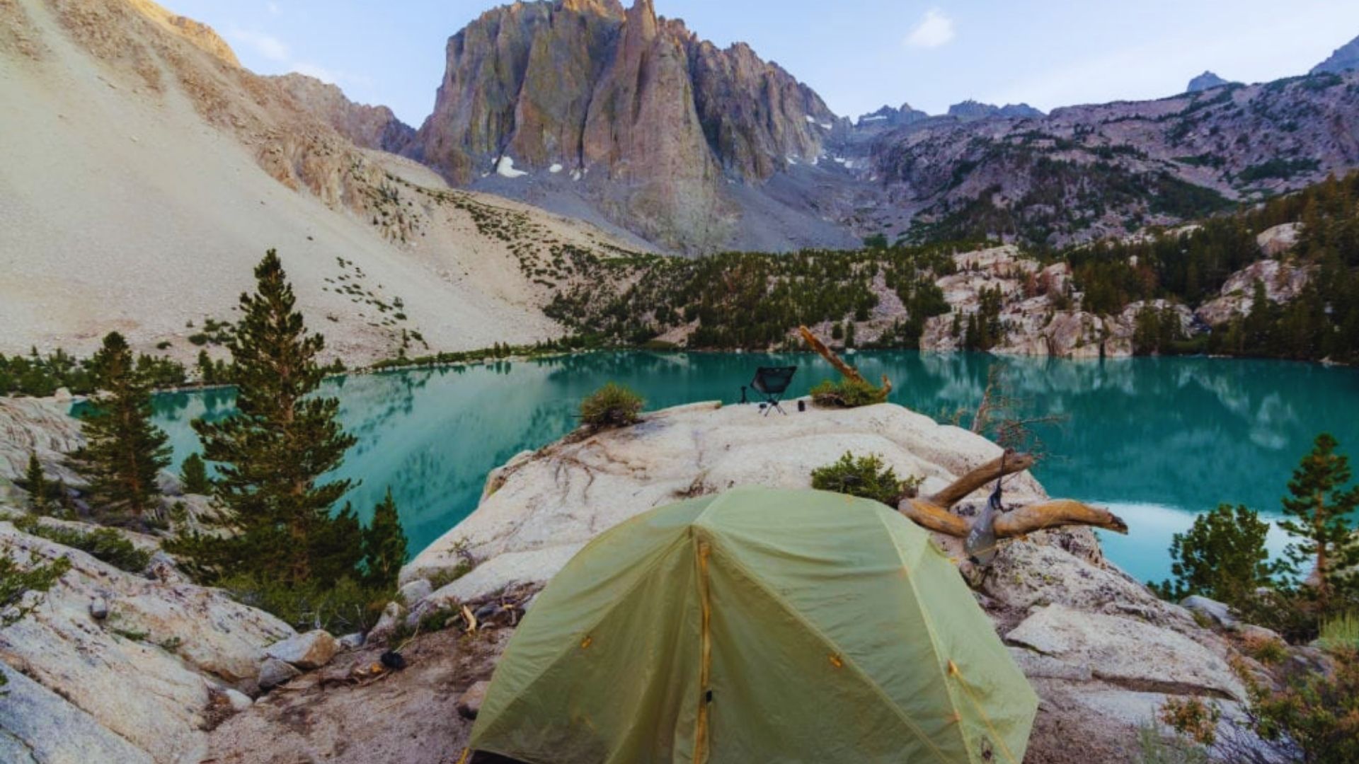 A serene mountain campsite with a tent surrounded by tall trees and rocky peaks under a clear blue sky.
