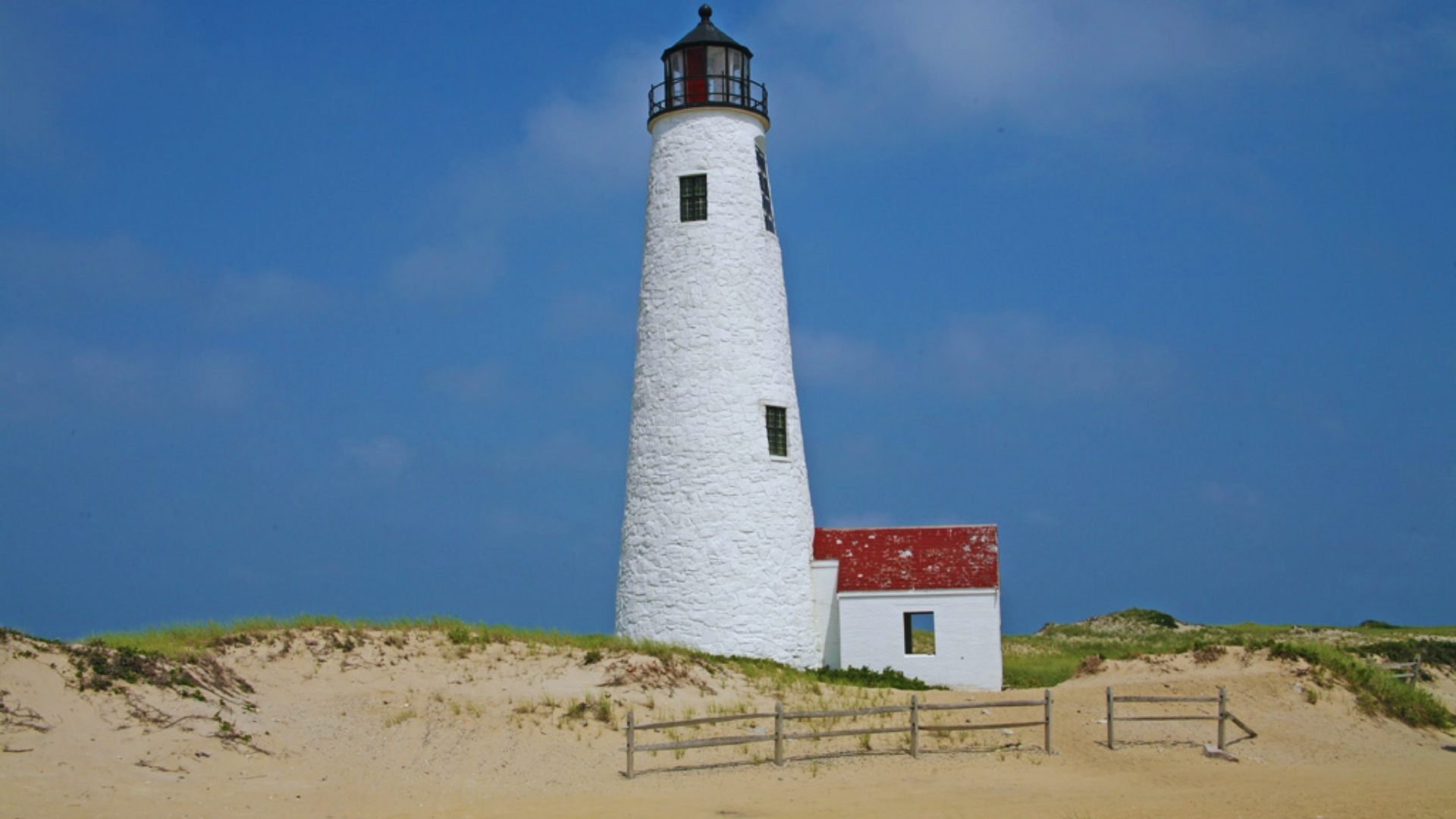 A white lighthouse stands on a sandy beach, overlooking the ocean under a clear blue sky.