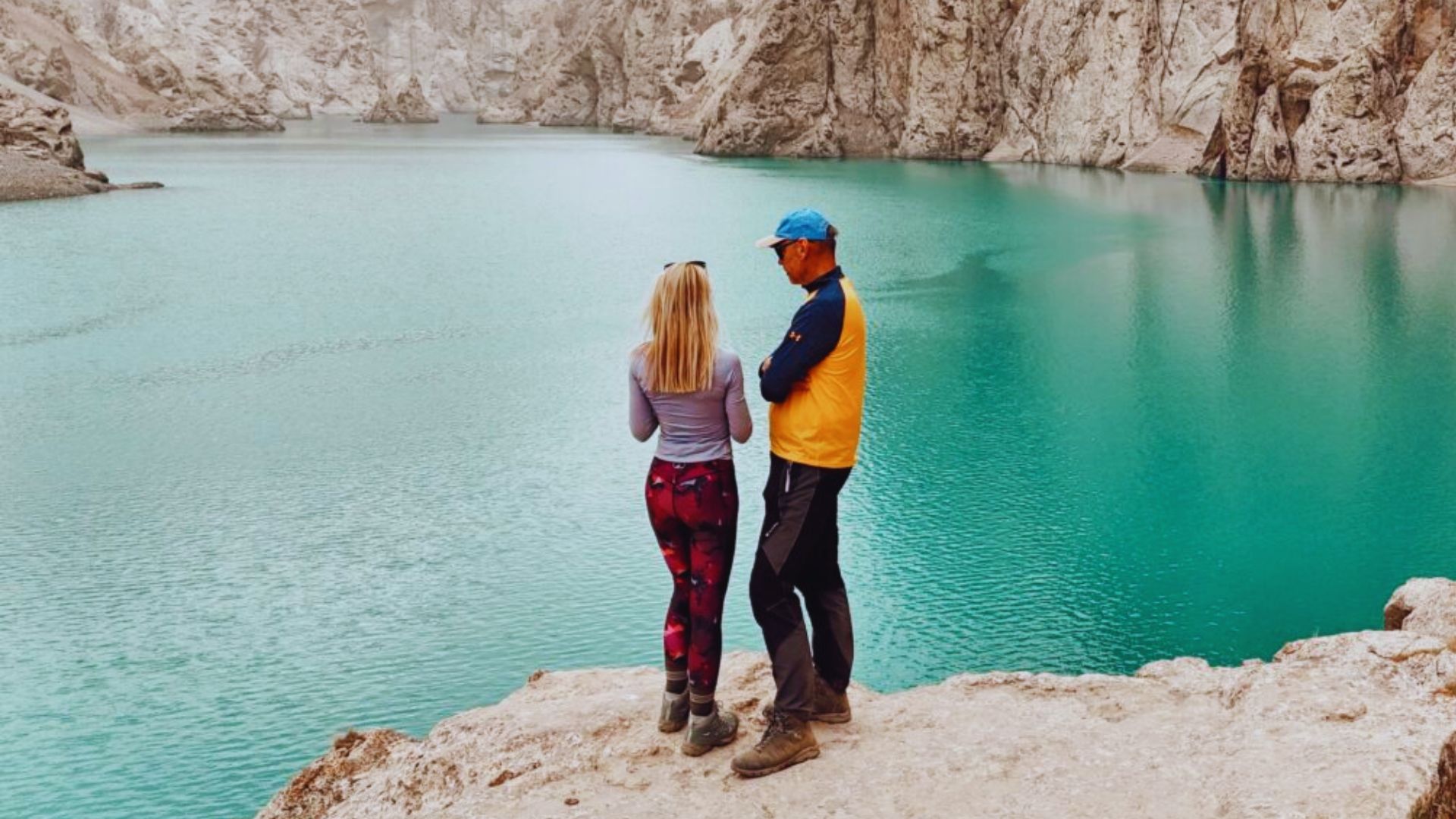 A couple stands on a cliff edge, admiring a picturesque lake below, with vibrant nature surrounding them.
