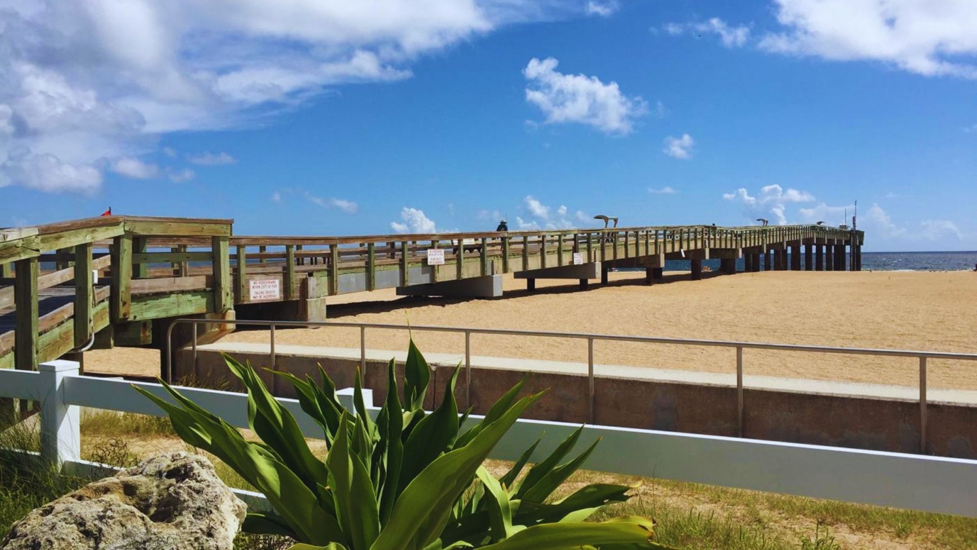 A long wooden pier extends over a sandy beach, with gentle waves lapping at the shore.