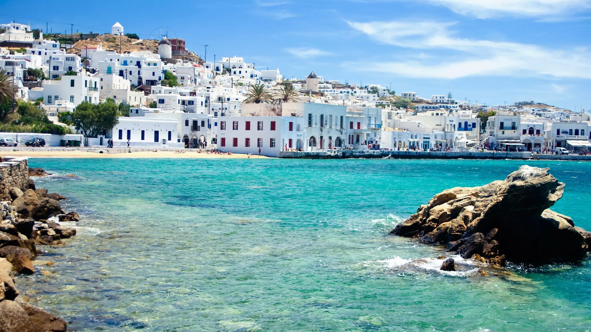 A scenic view of Mykonos beach in Greece, featuring golden sand and clear blue waters under a bright sky.