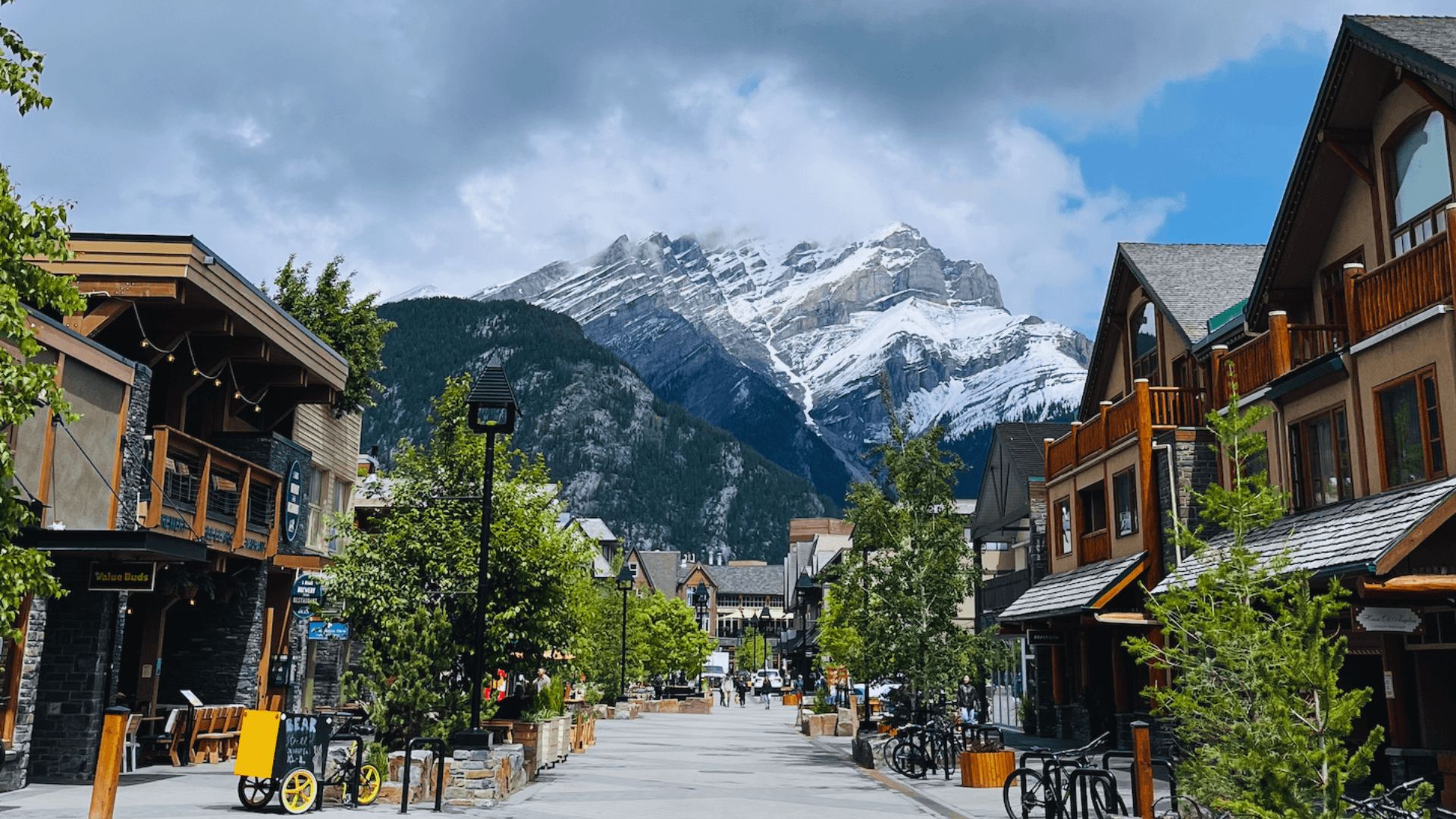 A city street featuring various buildings, with towering mountains visible in the distance.
