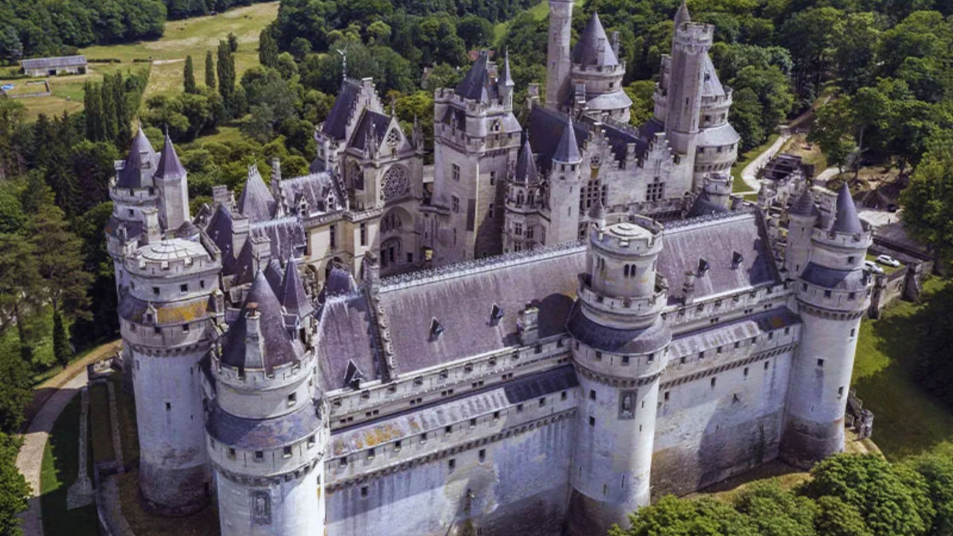 Aerial view of a grand medieval castle with tall towers and spires, surrounded by lush green forest. The scene conveys a sense of history and majesty.