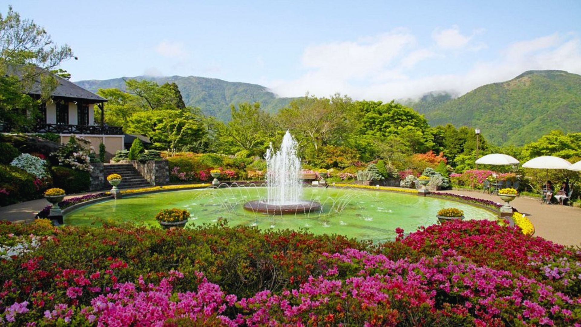 Lush garden with a central fountain surrounded by vibrant flowers, trees, and distant mountains. A serene path leads to shaded seating areas under umbrellas.