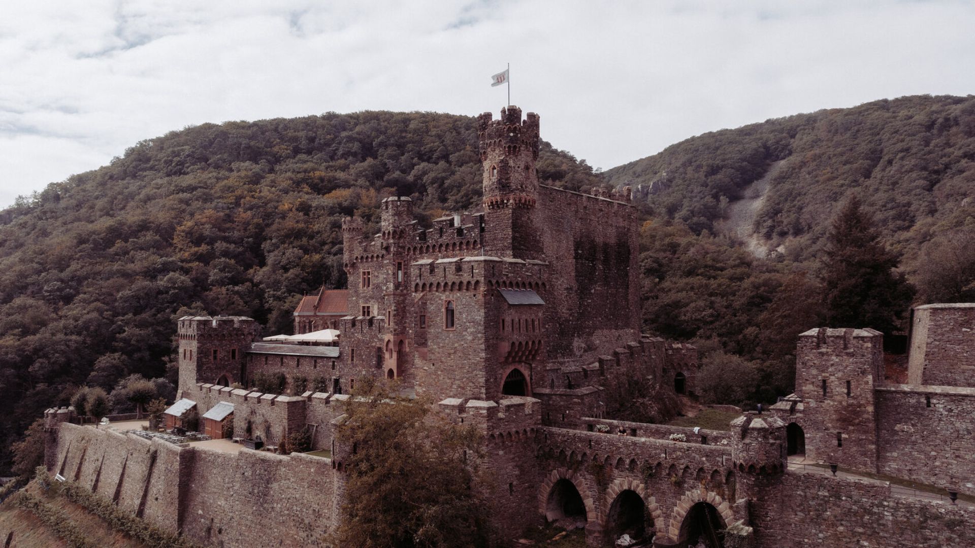 A medieval castle stands majestically amidst lush, rolling hills. The stone structure features towers, battlements, and an arched bridge, exuding a sense of historical grandeur.