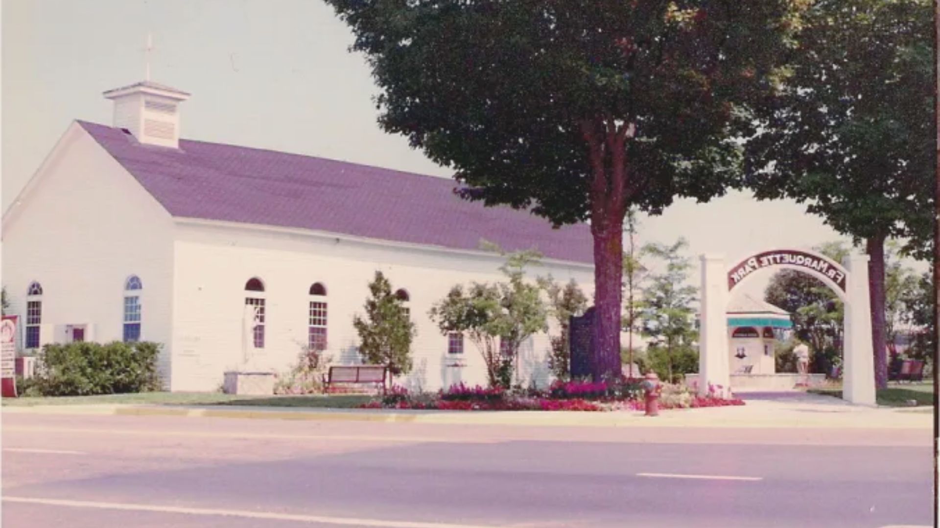 A quaint white chapel with a steeple and arched windows stands beside a large tree. A sign reads "Frankenmuth Park," and vibrant flowers line the pathway.