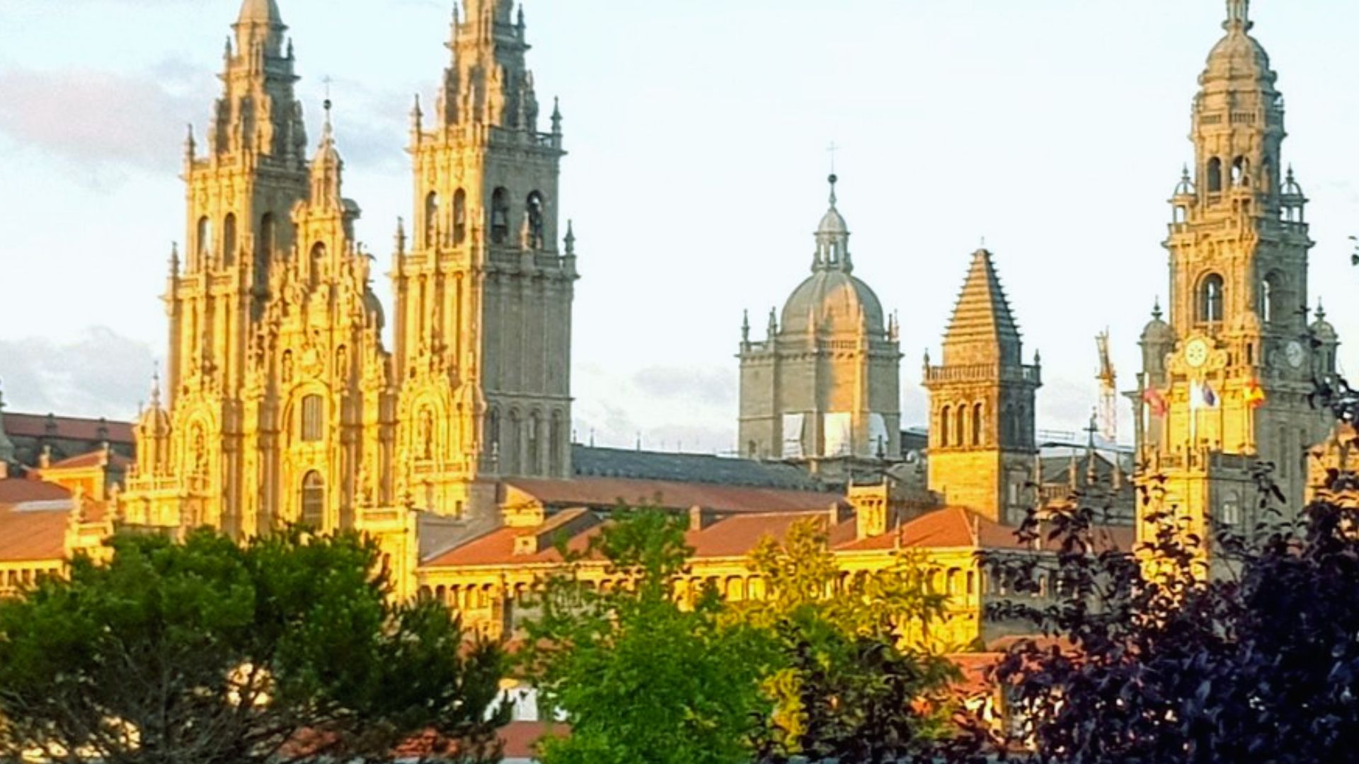 Sunlit view of a historic cathedral with intricate spires and domes, set against a partly cloudy sky, surrounded by lush greenery. The tone is serene.