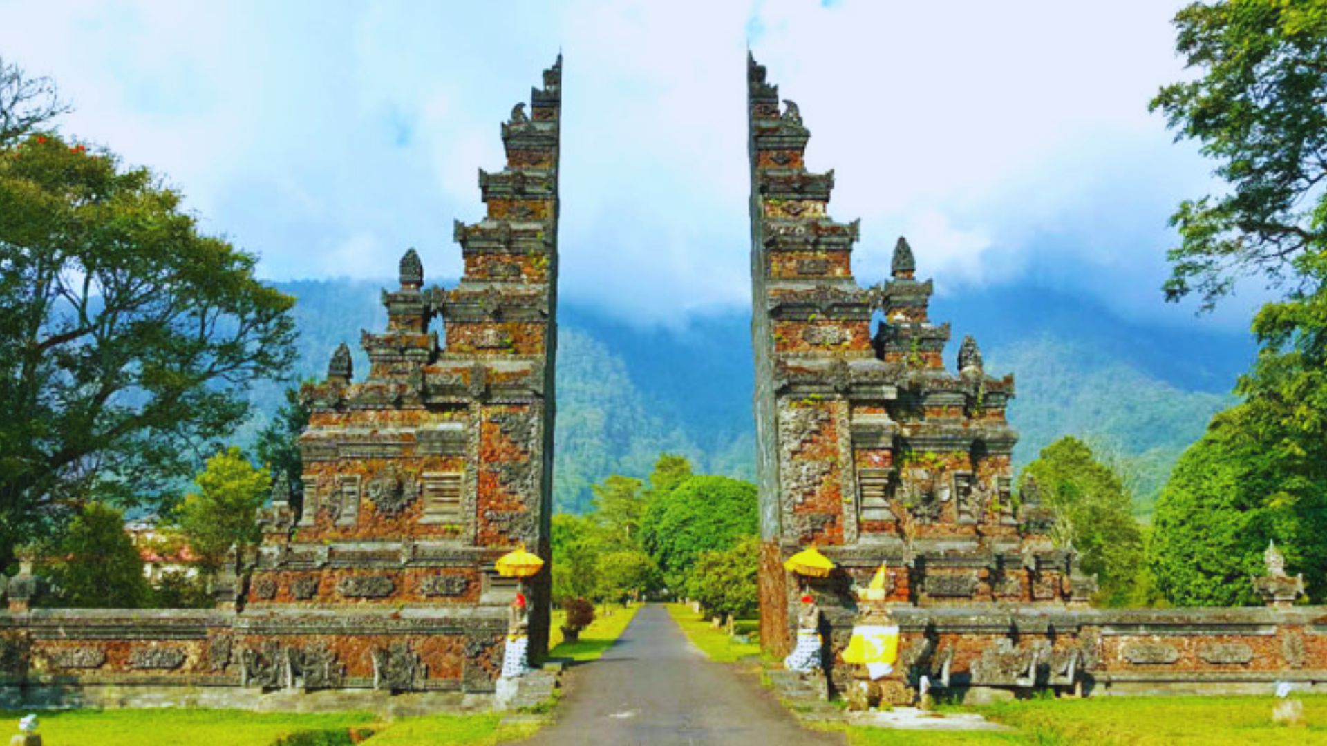 Ancient stone gate, split symmetrically, with intricate carvings. Flanked by lush trees under a bright sky, leading to a distant, misty mountain.