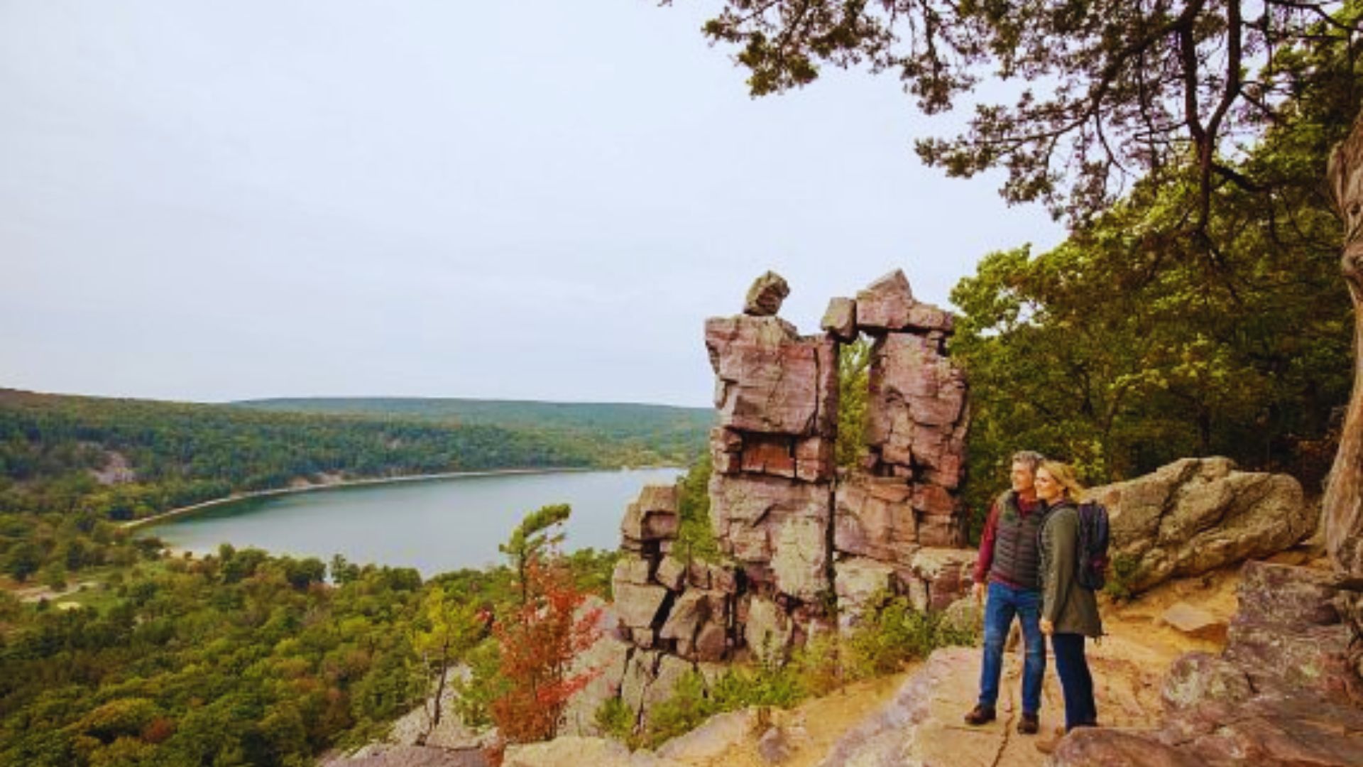 A couple stands on a rocky cliff overlooking a scenic view of a forest and a lake under a cloudy sky. The scene conveys a sense of adventure and tranquility.