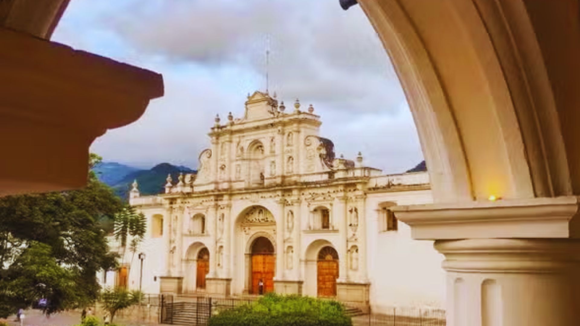 Historic white cathedral with ornate facade and three wooden doors, framed by an archway. Lush greenery and mountains under a cloudy sky. Peaceful ambiance.