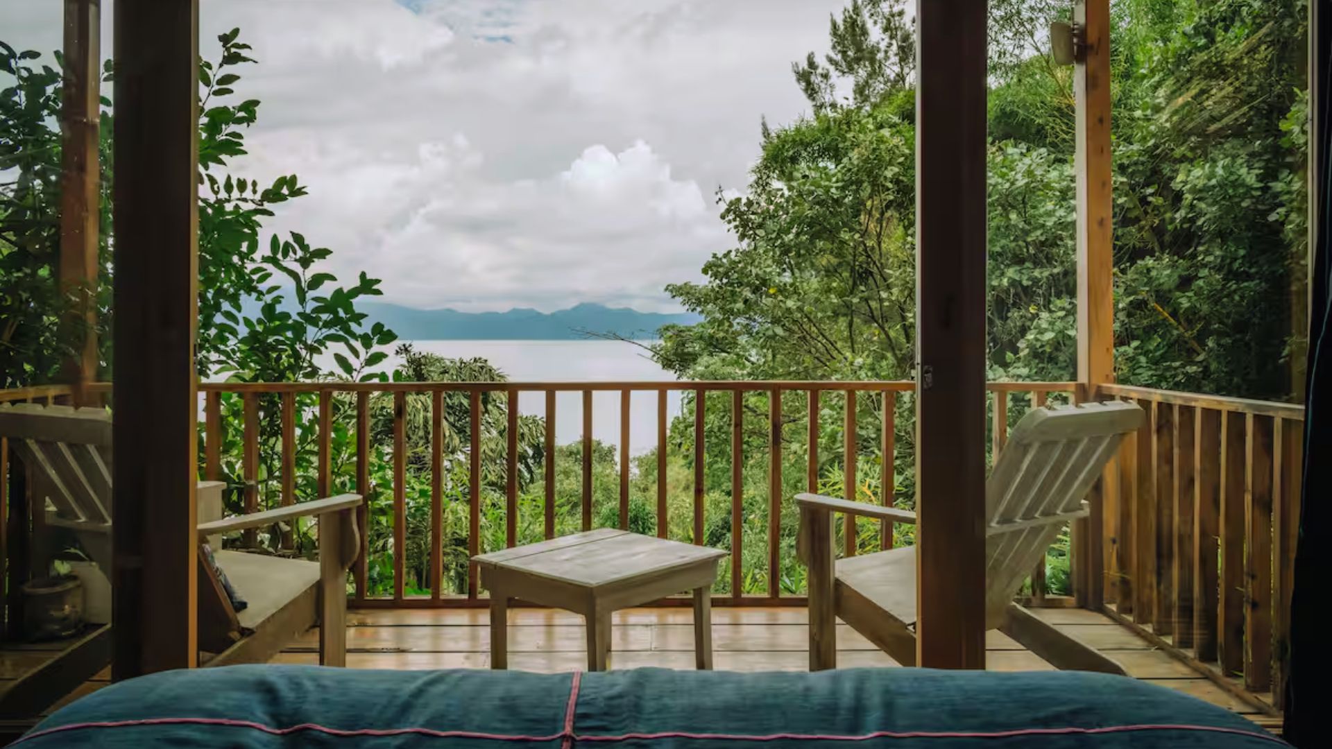 Wooden deck with two chairs and a table, surrounded by lush greenery. Overlooks a serene lake and mountains under a cloudy sky, conveying tranquility.