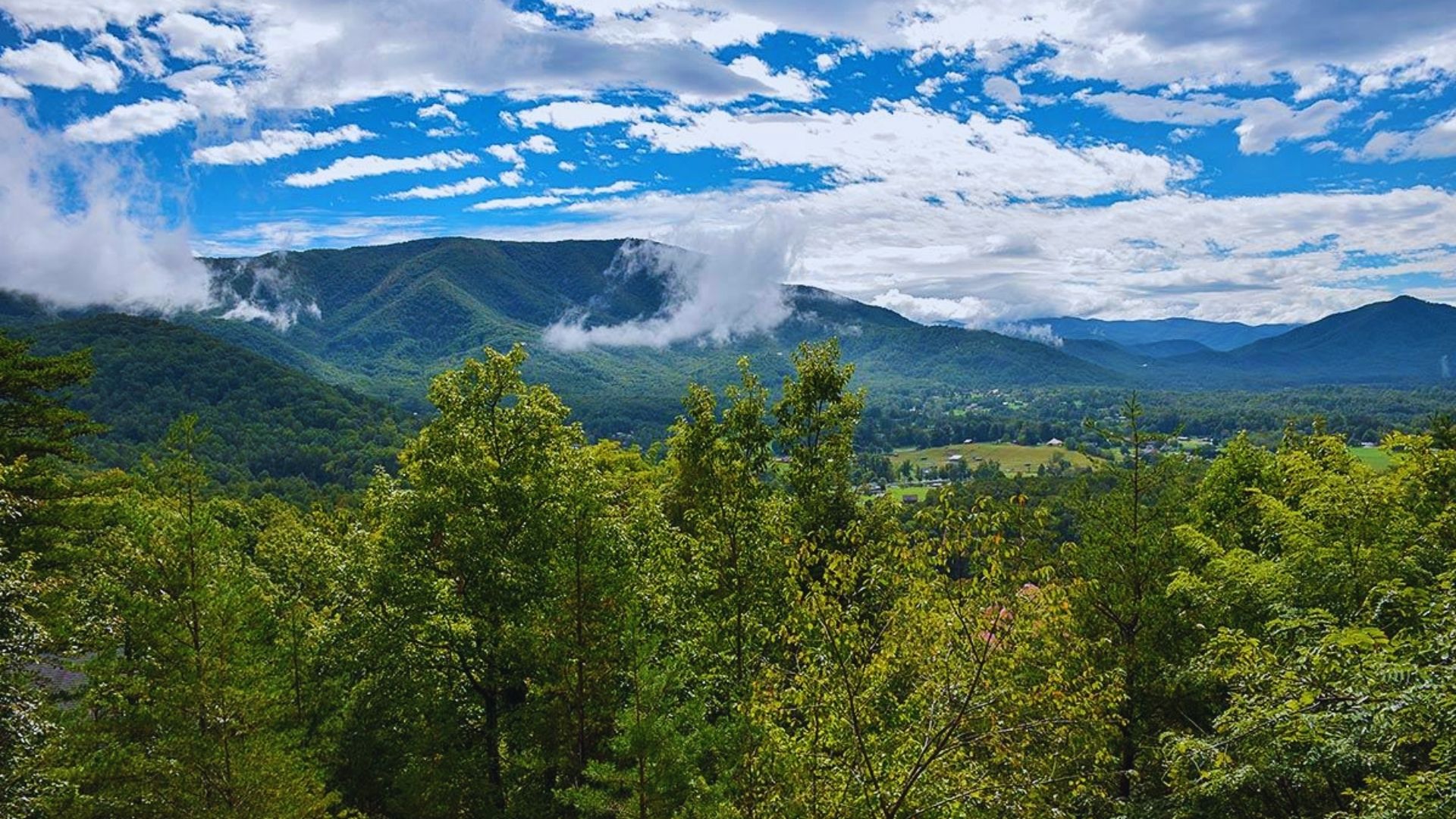 Lush green mountains under a bright blue sky with scattered clouds. Mist drifts through the valleys, creating a serene and peaceful landscape.