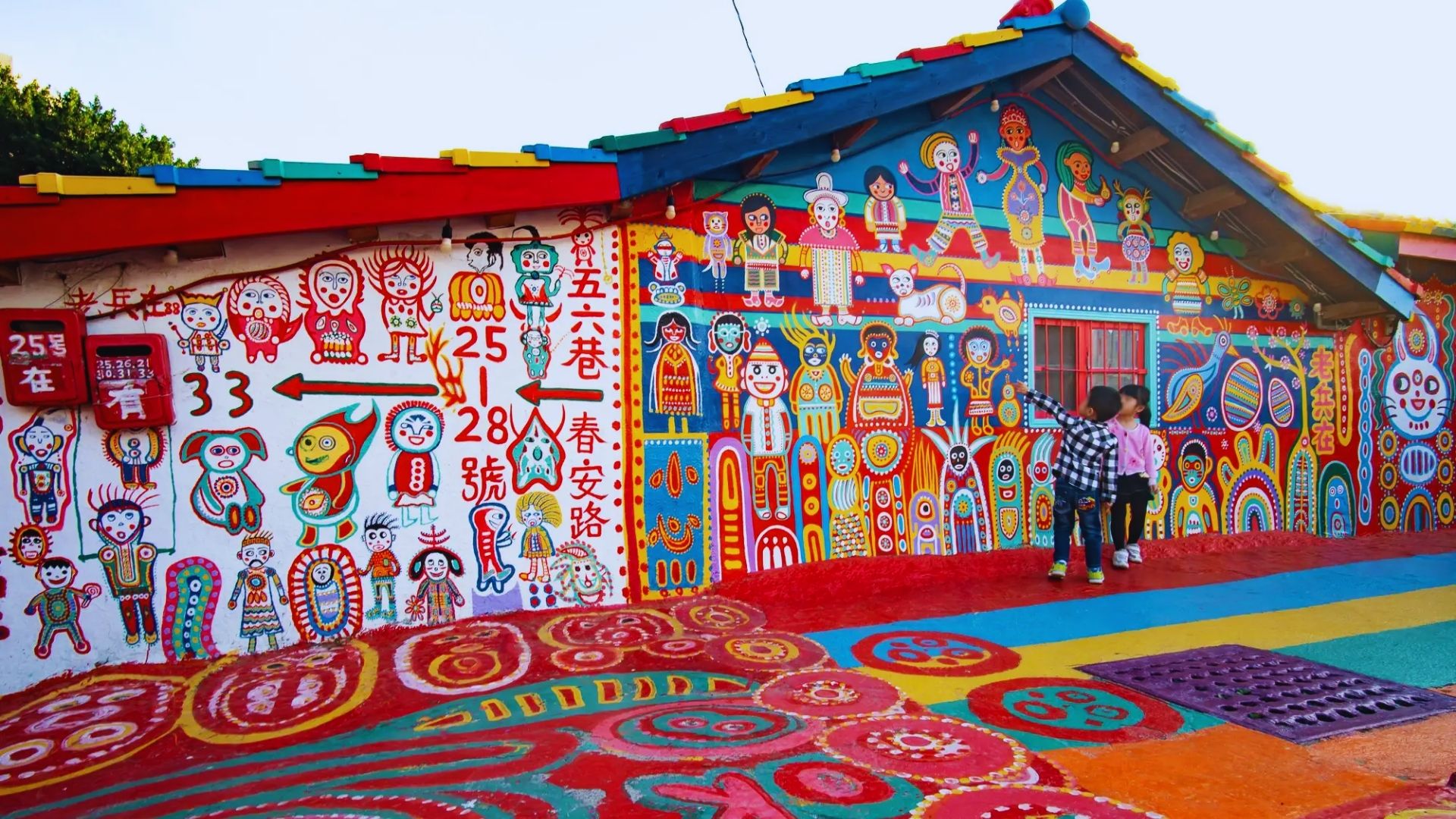 Brightly colored mural covers a house in vibrant reds, yellows, and blues. Painted figures and patterns, with two children admiring the artwork. Energetic and playful atmosphere.
