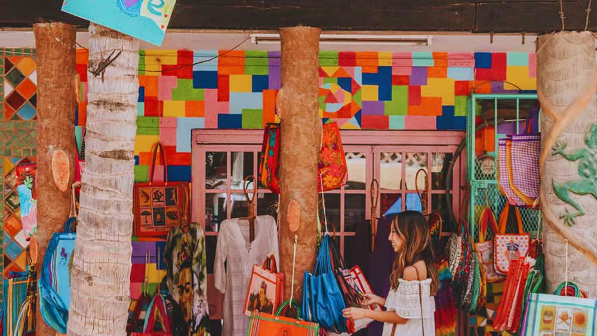 A woman in a white dress admires vibrant, colorful bags hanging outside a shop. The background is a lively mosaic wall, creating a cheerful, artistic atmosphere.