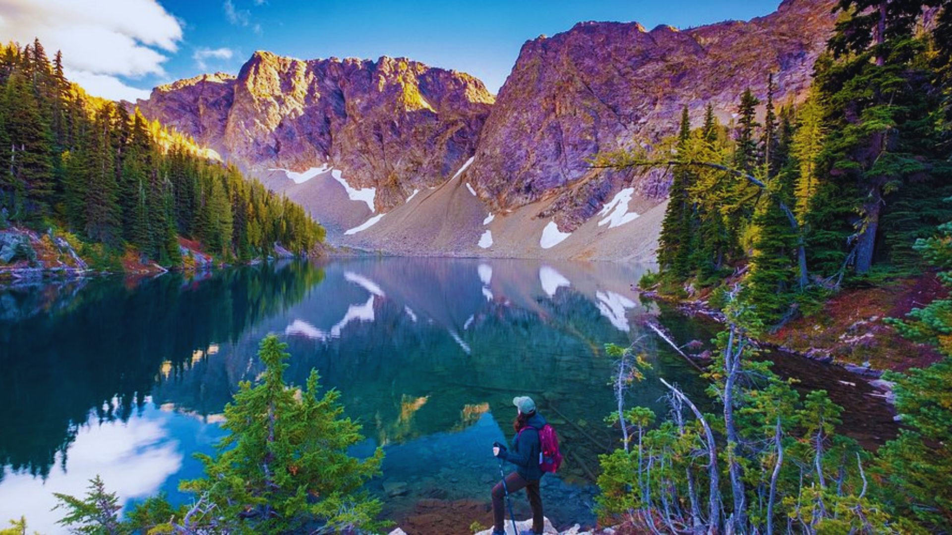 Hiker with a red backpack stands by a serene alpine lake reflecting rugged, sunlit mountains and pine trees under a vibrant sky, evoking adventure.