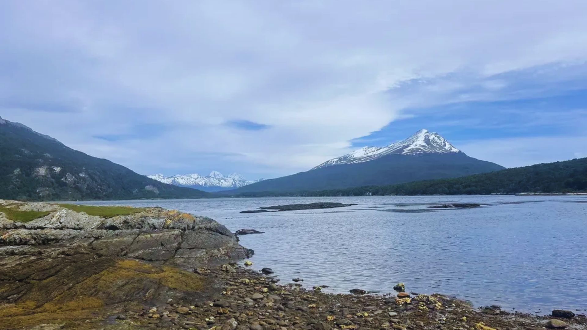A serene lake with rocky shores leads to a snow-capped mountain under a blue sky. The peaceful landscape conveys calmness and natural beauty.