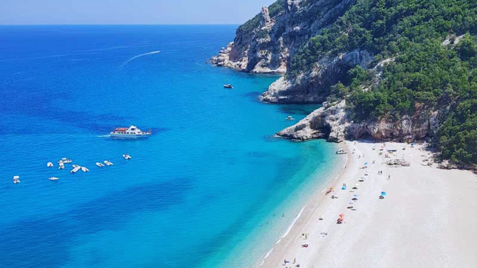 Aerial view of a picturesque beach with turquoise water and white sand. Small boats float near a rocky cliff, and colorful umbrellas dot the shore.