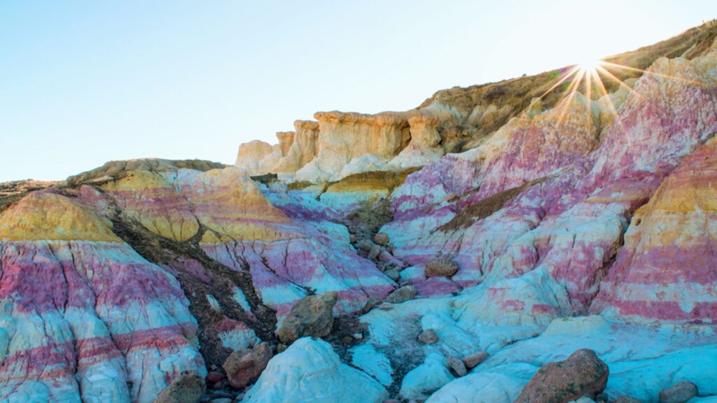 Paint Mines Interpretive Park: Complete Travel Guide Colorful rock formations in a desert, displaying layers of pink, purple, blue, and yellow under a bright sky. Sunlight glows over the ridge, creating a serene ambiance.