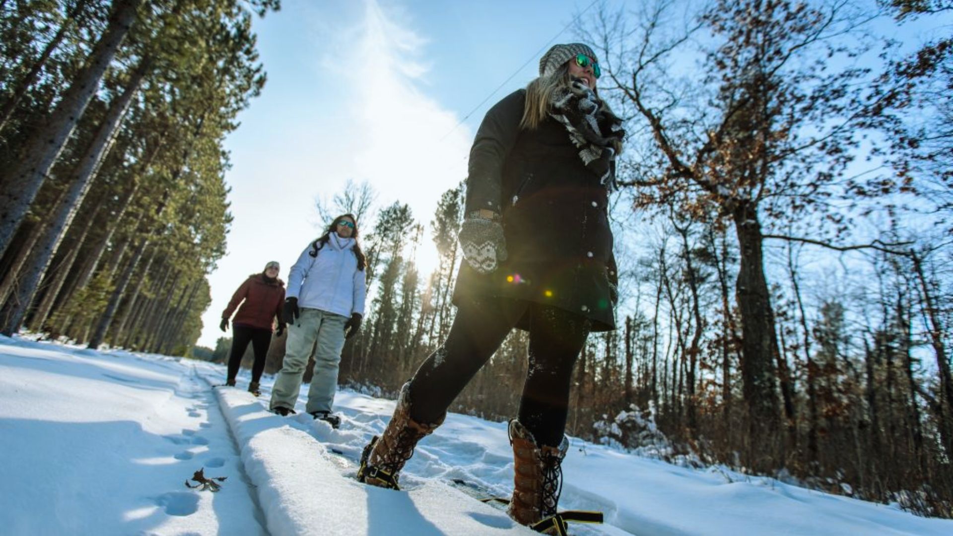Three people wearing winter clothing walk on a snowy path through a sunlit forest. The scene conveys a sense of adventure and enjoyment.