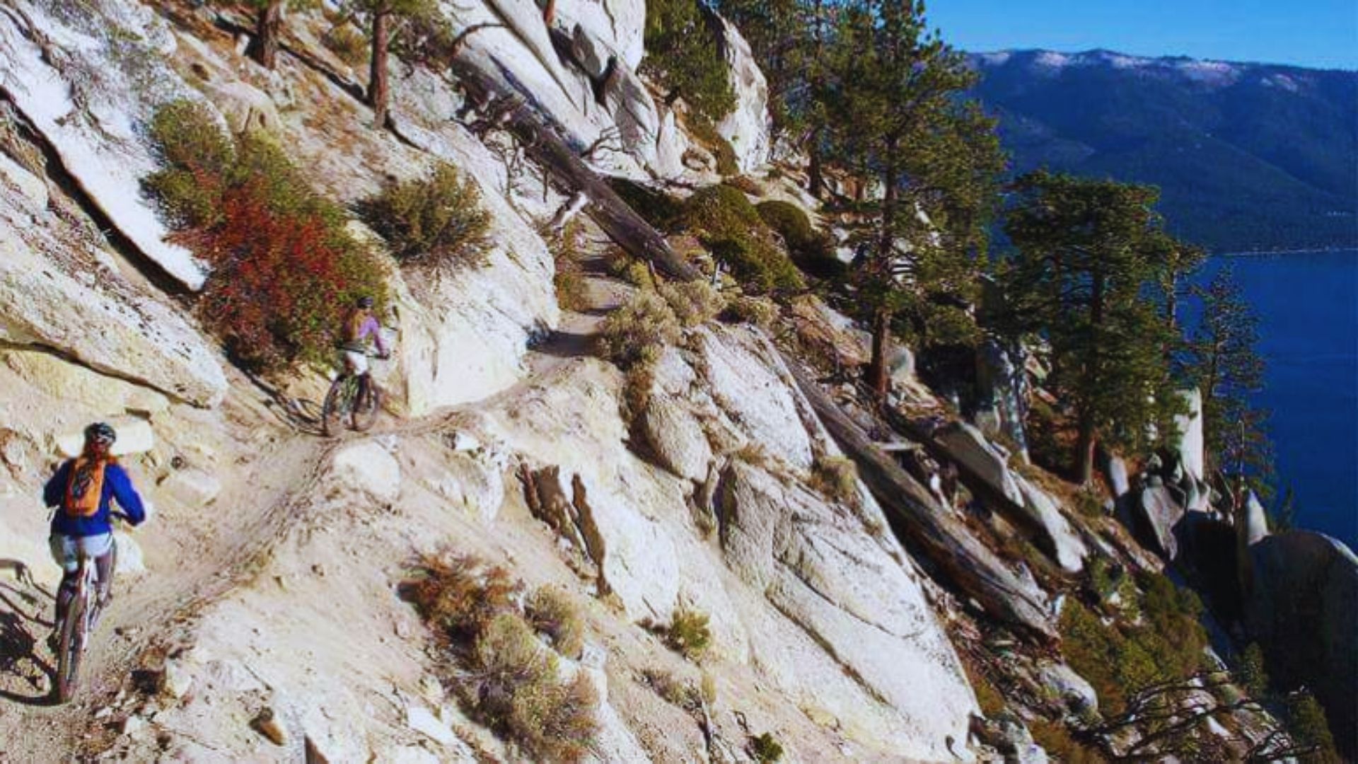 Two cyclists navigate a rocky trail alongside a body of water, enjoying the scenic outdoor landscape.