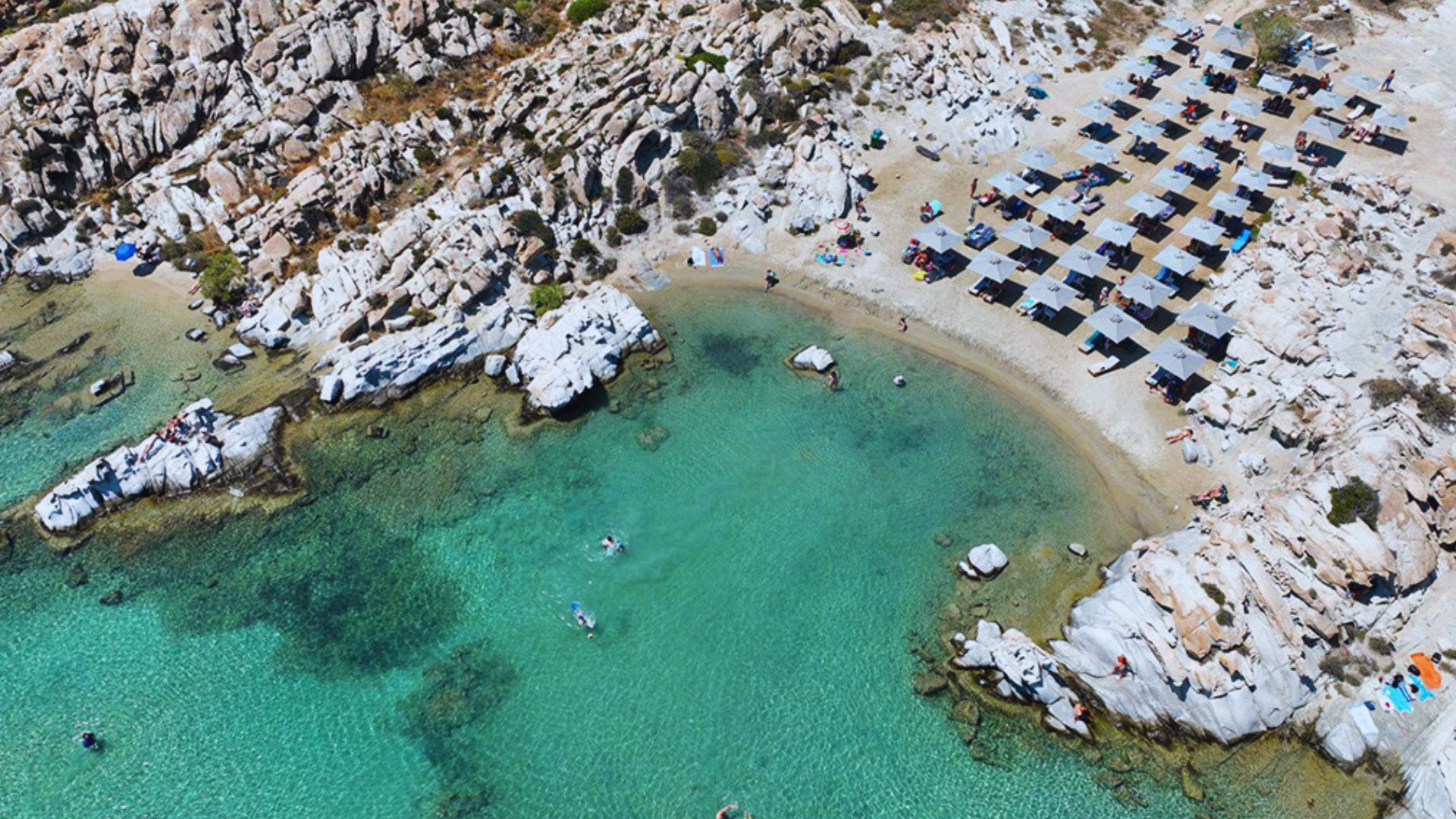 Aerial view of a rocky beach with clear turquoise water, sunbathers, and organized rows of sun loungers under umbrellas. The scene is serene and inviting.
